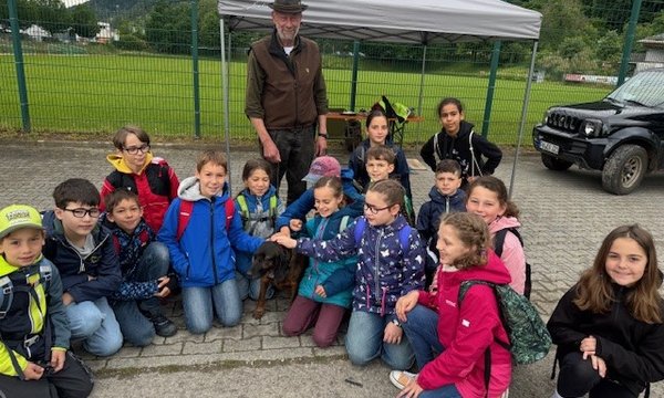 Eine Gruppe von Kindern kniet um einen Hund, während ein Mann daneben steht. Der Hintergrund zeigt eine Wiese mit einem Zaun und einem Auto.